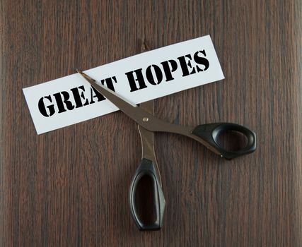 Scissors cutting the words "Great Hopes" written on a paper strip, over wooden background