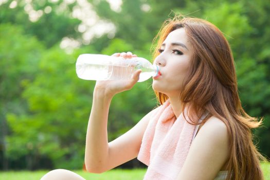 Woman sitting tired and drinking water after exercise. Within the lawn of park.