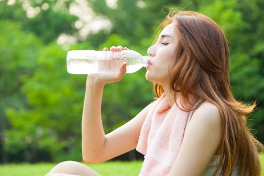 Woman sitting tired and drinking water after exercise. Within the lawn of park.