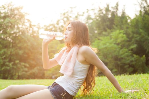 Woman sitting tired and drinking water after exercise. Within the lawn of park.