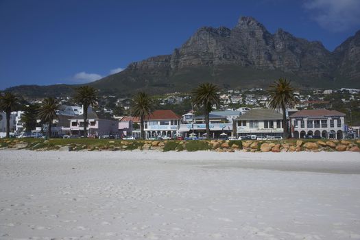 The Twelve Apostles mountains towering over the restaurants and bars lining the sandy beach at Camps Bay, Cape Town, South Africa