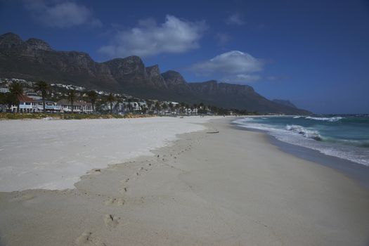 The Twelve Apostles mountains towering over the restaurants and bars lining the sandy beach at Camps Bay, Cape Town, South Africa.