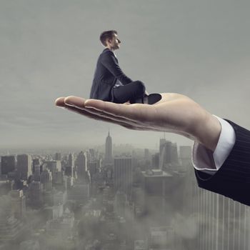 Businessman sitting on hand palm and looking away with cityscape and sky on background.