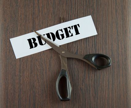 Scissors cutting the word "Budget" written on a paper strip, over wooden background