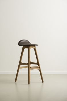 Moderrn high wooden bar stool with a black leather seat and footrests on a reflective floor in an empty room with a white wall, vertical format with copyspace in daylight