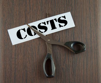 Scissors cutting the word "Costs" written on a paper strip, over wooden background