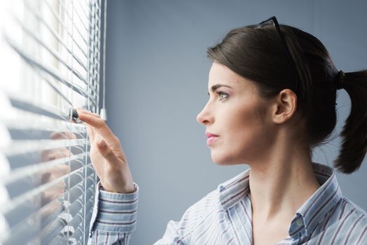 Young attractive woman peeking through blinds at window.
