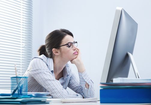 Bored office worker at desk staring at computer screen with hand on chin.