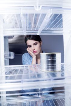 Young sad woman looking at one tin in her empty fridge.