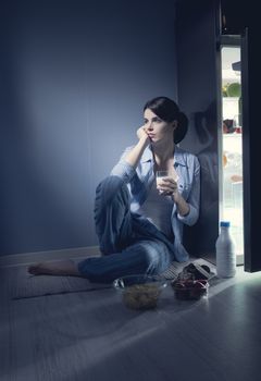 Sleepless sad woman sitting on kitchen floor having a glass of milk.