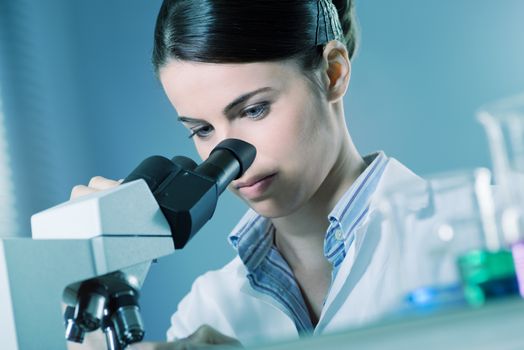 Young female researcher using microscope in the chemistry lab with laboratory glassware on foreground.