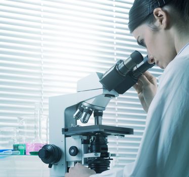 Young female researcher using microscope in the chemistry lab with laboratory glassware on background.