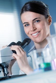 Female researcher using microscope and smiling at camera with beakers on foreground.