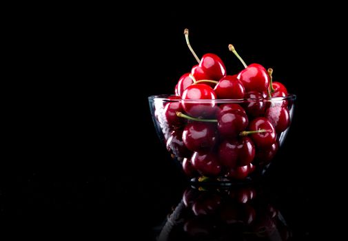 Juicy shiny cherries in a glass bowl on black background.