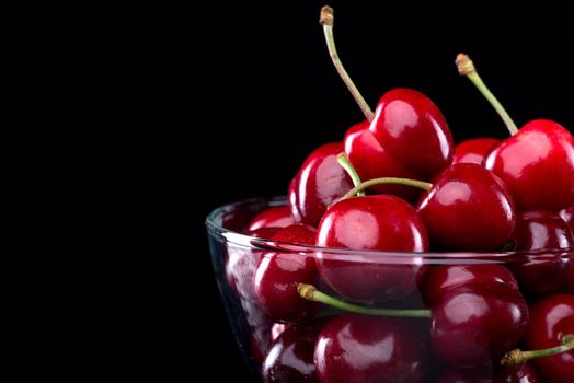 Juicy shiny cherries in a glass bowl on black background.