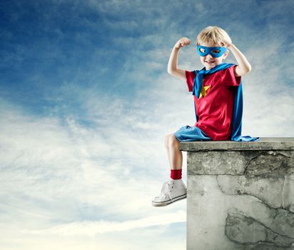 Cute little superhero boy with raised fists and blue sky on background.