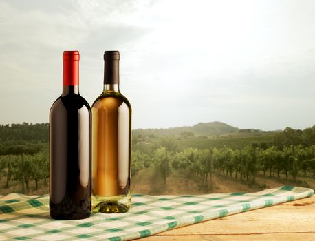 Red and white wine bottles standing on checked tablecloth and vineyard on background.