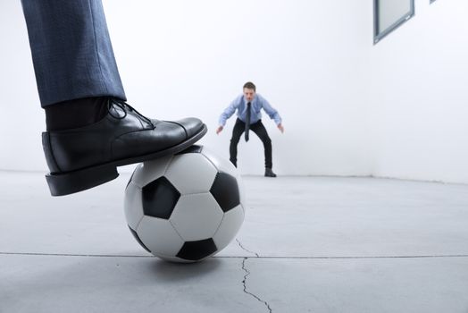 Businessmen playing soccer in an empty room, ball and foot close up with goalkeeper on background.