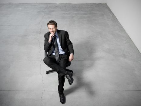 Smiling businessman sitting on an office chair with hand on chin against concrete floor background.