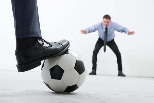 Businessmen playing soccer in an empty room, ball and foot close up with goalkeeper on background.