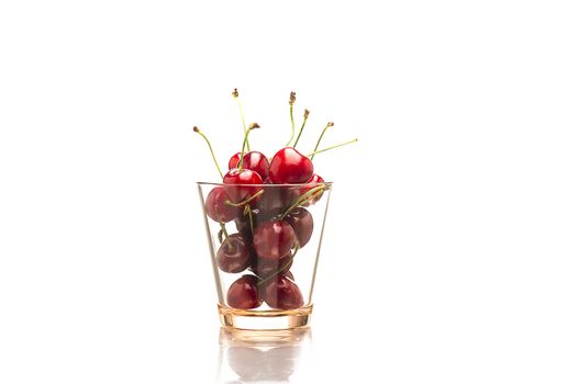 Horizontal close-up of a transparent glass full of red fresh cherries, natural source of vitamin C and minerals, with reflection on white