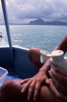 Sunbather on boat with far leg in focus applying sun lotion, against an Indian Ocean island background
