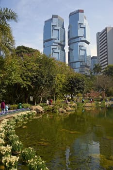 People walking around a lake in Hong Kong Park surrounded by tall office buildings.