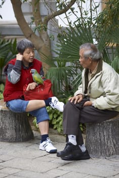 Woman with a parrot perched on her leg sitting talking to a man in the Yuen Po Street Bird Garden in Kowloon, Hong Kong.