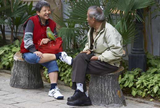 Woman with a parrot perched on her leg sitting talking to a man in the Yuen Po Street Bird Garden in Kowloon, Hong Kong.