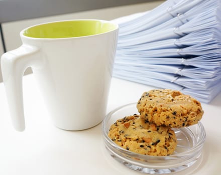 Multiple documents lay stacked on the desk.The front has a coffee cup and two pieces of cookie are placed in the dish.                             