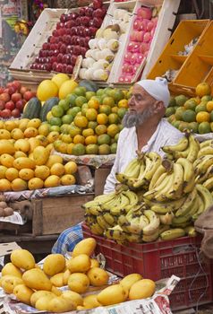 Old man with beard sitting amongst fruit and vegetables on a market stall in Old Delhi, India.