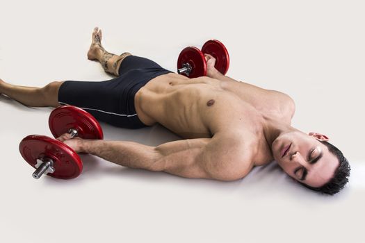 Exhausted young male bodybuilder resting on floor with dumbbells in hands
