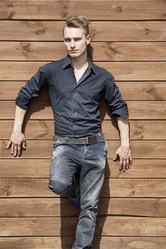 Handsome blond young man standing outside against wood wall looking at camera