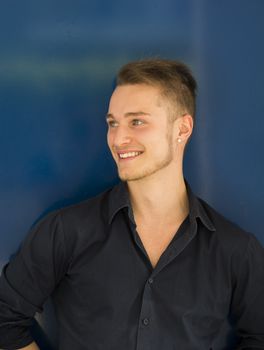 Attractive blond, blue eyed young man smiling and looking away, against blue metal wall