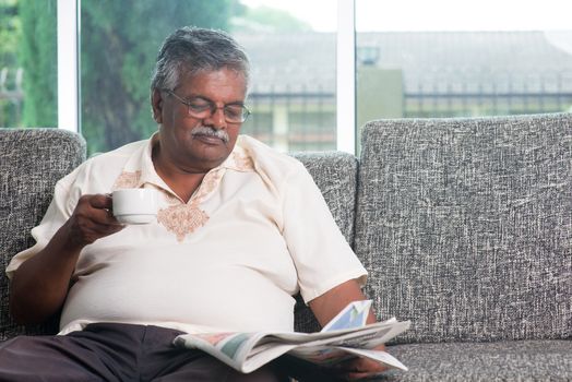 Asian Indian senior adult drinking coffee while reading news paper sitting on sofa at home in morning, elderly retirement indoor living lifestyle.