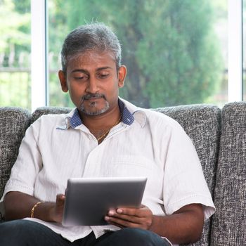 Portrait of middle aged Indian man reading on digital tablet computer at home.