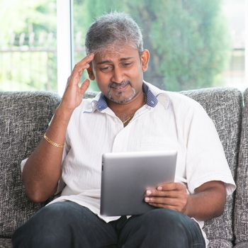 Portrait of middle aged Indian man headache while reading on digital tablet computer at home.