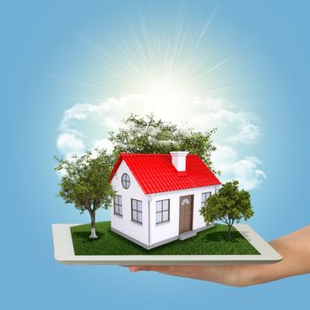 Hands holding a tablet pc and small house with land. Sky and sun on background
