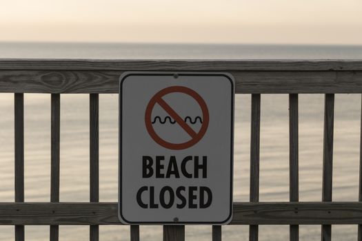 Beach is closed sign posted on wooden pier. Calm waters in the background.
