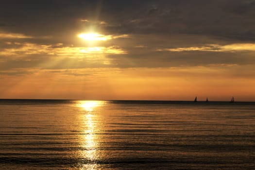 Orange sparkling sun shines through the clouds and reflects onto calm water. Sailboats in the far distance. 