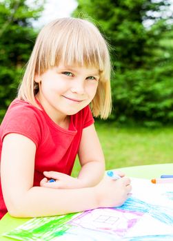Little girl sitting at table drawing a house outdoors