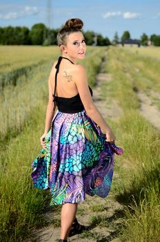 Young girl walks on dirt-track surrounded by green fields with blue sky as background. Female model from Poland wears colorful dress.