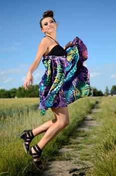 Outdoor photo of happy female model jumping. Young Polish model wearing colorful dress. Green fields and blue sky as background.