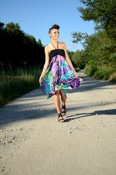 Young girl walks on dirt-track surrounded by green fields with blue sky as background. Female model from Poland wears colorful dress.