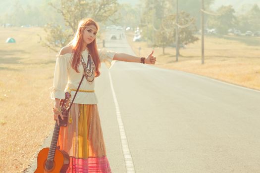 Hippie girl with guitar hitchhiking on countryside road