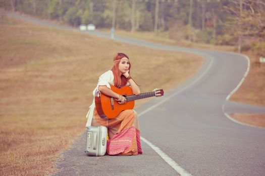 Hippie young woman with guitar and backpack sitting on a countryside road