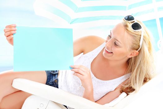 Beautiful blonde woman sitting on a lounger beach holding a blue sign with space for text.