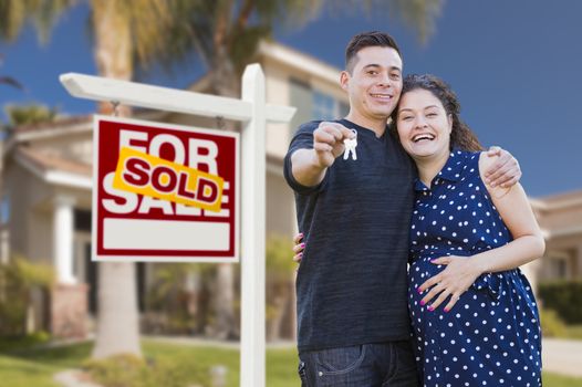 Young Happy Pregnant Hispanic Young Couple with House Keys in Front of Their New Home and Sold For Sale Real Estate Sign.