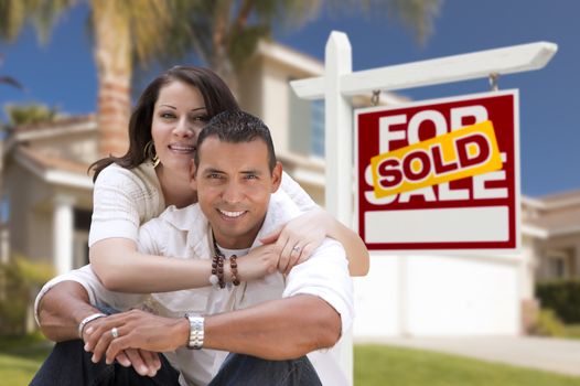 Young Happy Hispanic Young Couple in Front of Their New Home and Sold For Sale Real Estate Sign.