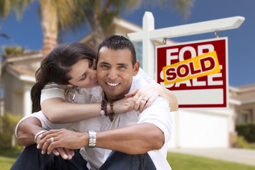 Young Happy Hispanic Young Couple in Front of Their New Home and Sold For Sale Real Estate Sign.